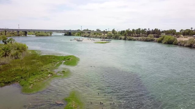 aerial footage of the silky green waters of the Colorado River with people in boats on the water surrounded by lush green palm trees and grass with blue sky at Quechan Park in Blythe California USA