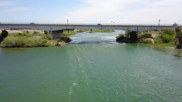 aerial footage of the silky green waters of the Colorado River with a metal bridge over with water with cars driving surrounded by lush green trees and plants with blue sky at Quechan Park in Blythe