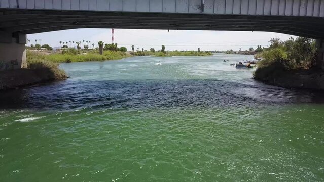 aerial footage of the green waters of the Colorado river with people riding jet skis near a metal bridge with cars surrounded by lush green palm trees and plants at at Quechan Park in Blythe