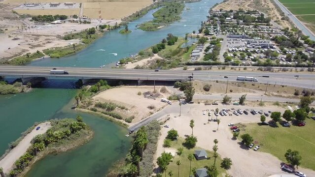 aerial footage of the silky green waters of the Colorado river with people riding jet skis and sailing boats and a street with cars driving and lush green palm trees and grass at Quechan Park