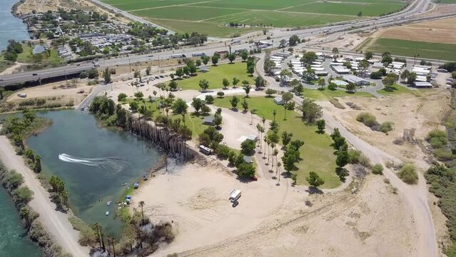 aerial footage of the silky green waters of the Colorado river with people riding jet skis and sailing boats and a street with cars driving and lush green palm trees and grass at Quechan Park