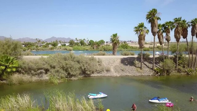 aerial footage of the silky green waters of the Colorado River with people in boats on the water surrounded by lush green palm trees and grass with blue sky at Quechan Park in Blythe California USA