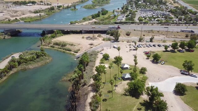 aerial footage of the silky green waters of the Colorado river with people riding jet skis and sailing boats and a street with cars driving and lush green palm trees and grass at Quechan Park