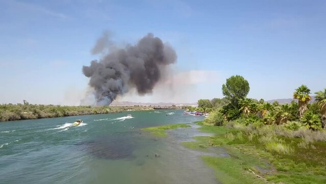 aerial footage of the green waters of the Colorado River with people in boats and smoke from a brush fire on the banks of the river with blue sky at Quechan Park in Blythe California USA
