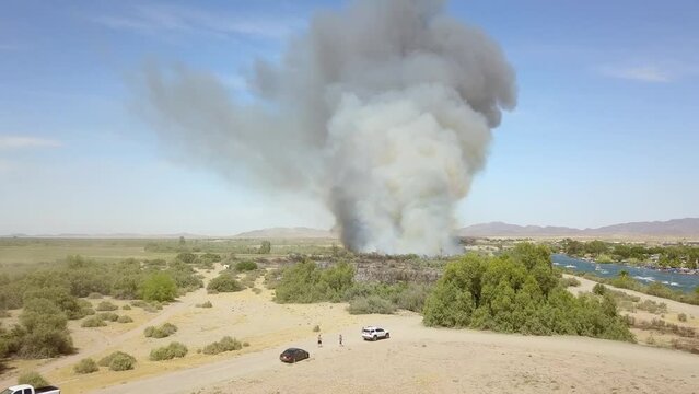 aerial footage of a brush fire along the silky green waters of the Colorado River surrounded by dry brush and lush green trees and plants with boats on the water and cars driving with blue sky