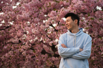 smiling Asian young man arm crossing, side face looking away. Blur pink sakura tree background