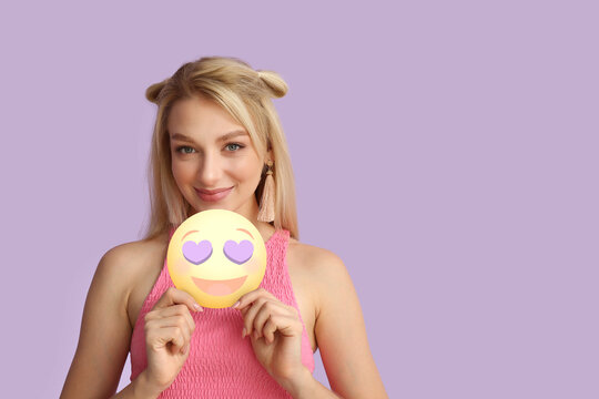 Pretty young woman holding happy emoticon with heart-shaped eyes on lilac background