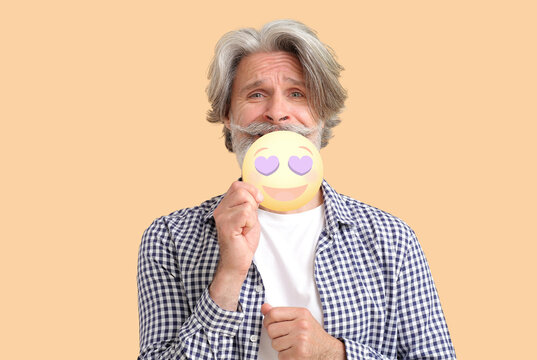 Senior man holding happy emoticon with heart-shaped eyes on color background