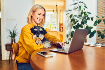 Cheerful mature woman using laptop sitting with dog at a table working remotely