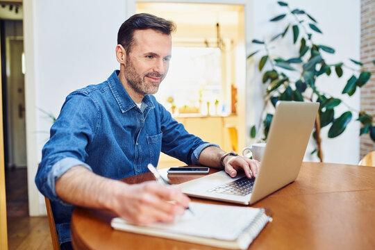 Mid adult man using laptop writing notes while at table working remotely