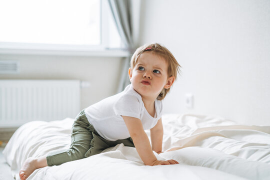 Cute upset baby girl angry child in home clothes sitting on bed at home