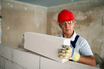 Bricklayer or mason lays bricks to construct wall of autoclaved aerated concrete blocks. Brickwork worker contractor doing precise masonry of foam concrete with spirit and laser levels.