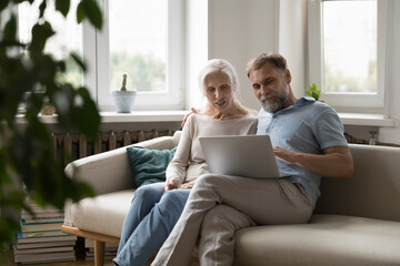 Happy focused elder 60s couple sitting on couch at home, using laptop, making video call, talking to family together, shopping online, using online app, internet payment services