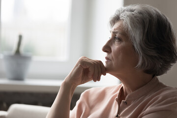 Frustrated sad mature grey haired 50s woman thinking over bad news, sitting on couch at home, feeling depressed, mourning, going through loss, grief, health problems, mental disorder, cancer disease