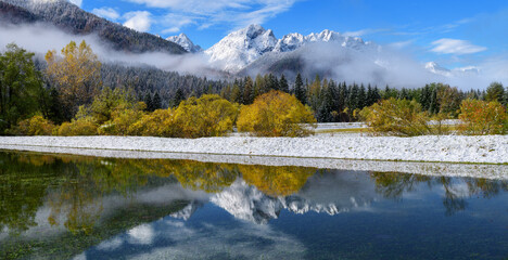 When autumn meets winter at the lake with perfect reflections in a calm water. Autumn trees with fresh snow and fog. 