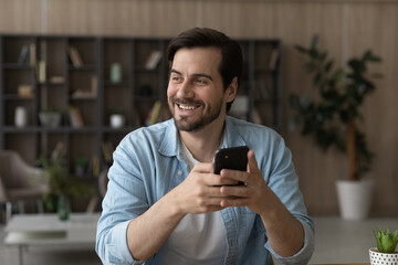 Dreamy and inspired. Happy casual young man holding smartphone looking at distance thinking about...