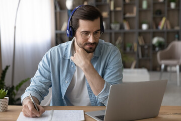Busy at home office. Focused male student in wireless headphones look on pc screen do homework write training material translation to english. Young man improve professional skills at virtual workshop