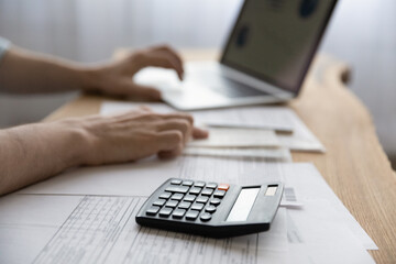 Bookkeeping. Close up of young man accountant financial analyst work with papers invoices statements use computer to declare taxes pay bills on distance. Focus on electronic calculator lying on desk
