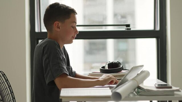 Caucasian boy sitting at table with workbook and looking at tablet screen while attentively listening to teacher online.