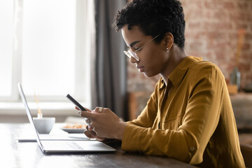 African woman sit at desk holds smartphone. Distracted from work on wireless computer, lead sms...