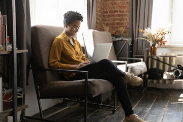 Young 25s African woman in glasses put computer on laps, work on laptop while sit on armchair in...