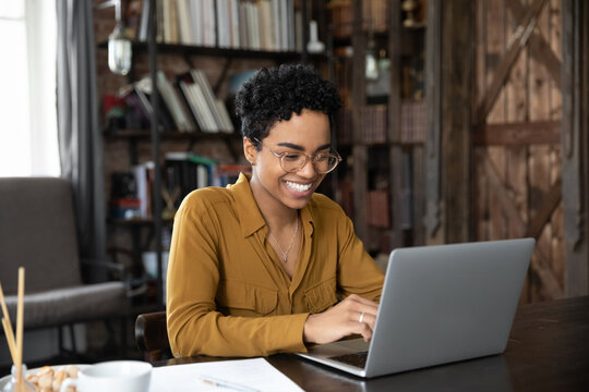 African woman sit at desk working on laptop, student studying makes assignment looks happy, feels satisfied. Workflow use modern tech, chat on-line, share messages with friends in social media concept - Powered by Adobe