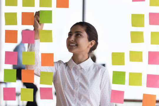 Attractive young Indian female employee jotting information and reminders on colorful sticky notes attached on glass wall in office. Create to-do list, develop fresh start up business ideas concept
