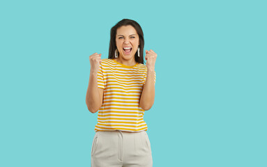 Beautiful woman receives good news, wins competition, gets prize, celebrates success and victory. Happy excited euphoric lady in striped tee isolated on blue background fist pumping and shouting Yes