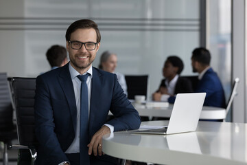 Handsome millennial smiling businessman in elegant formal suit sit at desk with laptop staring at cam, diverse businesspeople on background. Leadership, careerism, business application usage concept