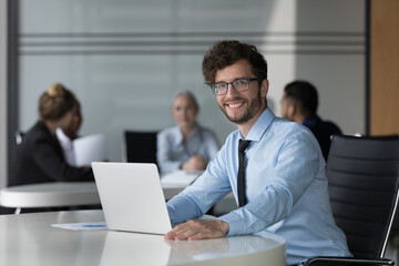 Young office employee, confident businessman sit at desk with laptop smile look at camera, feels satisfied by career growth in company, colleagues on background. Tech, promoted worker portrait concept