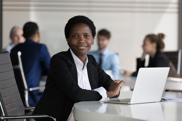 Successful African businesswoman sit at desk with laptop look at camera feel satisfied by career in international company, make online task use corporate software. Confident lady boss portrait concept