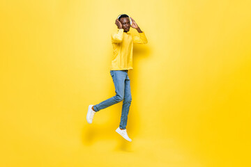 Fototapeta na wymiar Young African man listening to music on headphones and jumping in isolated yellow studio background