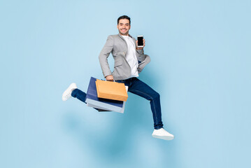 Young smiling Caucasian man holding shopping bags and mobile phone while jumping in light blue...