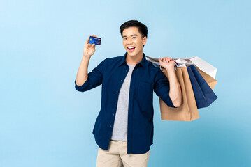 Smiling Asian tourist man carrying shopping bags and holding credit card in isolated light blue...