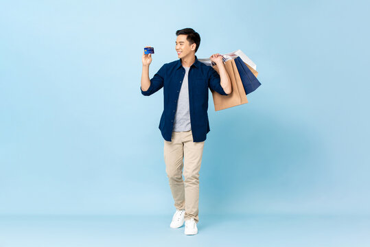 Full length portrait of handsome Asian tourist man carrying shopping bags and holding credit card in isolated light blue color studio background
