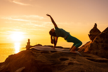 Urdhva Dhanurasana, a woman doing meditation and yoga exercises on a rock at sunset next to a lighthouse in the sea, healthy and naturist life
