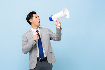 Young handsome Asian man in formal business suit holding megaphone in isolated light blue color...
