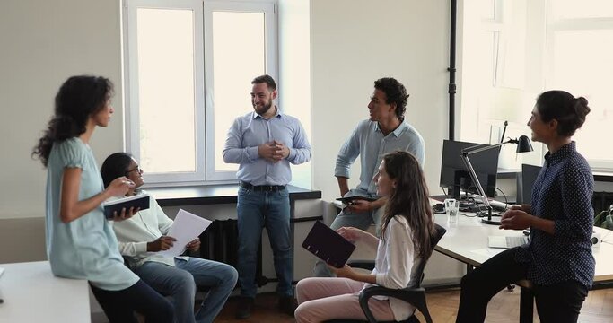 Young male international company department leader talk to smiling multiracial colleagues on brief meeting at workplace share plans with staff. Diverse teammates friends discuss job issues on seminar