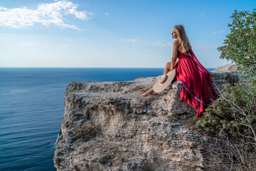 A girl with flowing hair in a long red dress sits on a rock above the sea. The stone can be seen in the sea.