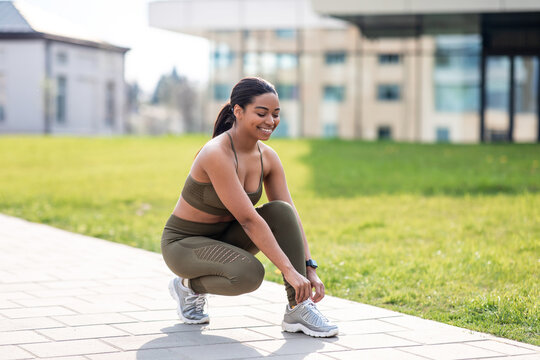 Cheerful young African American woman tying laces on her sneakers before training, jogging at city park, empty space
