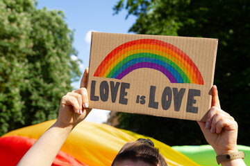 Woman holding placard sign Love is Love with rainbow, symbol of LGBT community. Giant flag in background. Pride Parade, equality march to support and celebrate LGBT+, LGBTQ Gay and lesbian equality. - obrazy, fototapety, plakaty