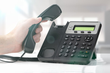 contact us. voip telephone for communication, contact us and customer service support. landline telephone device at office desk. hand dialing number and holding telephone receiver - obrazy, fototapety, plakaty