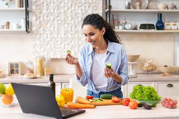 Happy latin woman cooking dinner and video calling on laptop, talking to webcamera, standing in kitchen at her apartment