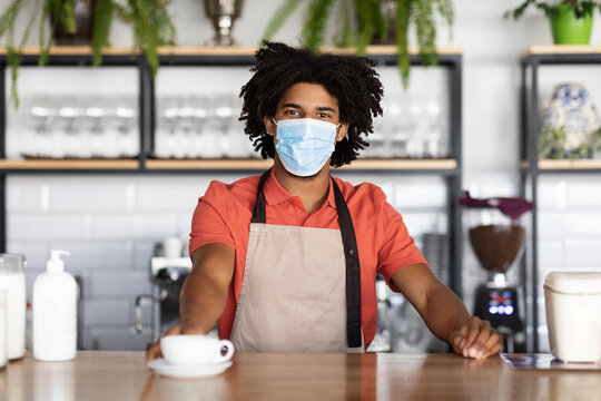 Smiling curly young black guy bartender in protective mask and apron gives cup of coffee to client in cafe