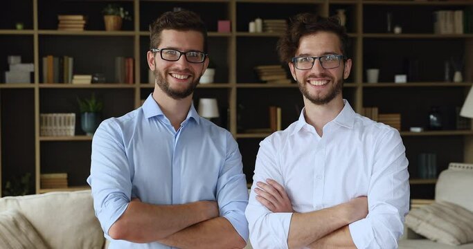 Two confident 35s male twins in glasses wear elegant shirts pose together standing in modern home with arms crossed on chest smile look at camera. Family bond, best friends brothers portrait concept