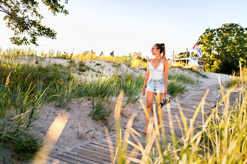 Summer sunset walk on beach boardwalk, green grass field and sand dunes. Outdoor nature portrait of happy pretty girl. Woman with trendy stylish vacation outfit. Fashion lifestyle. Weekend vibes.