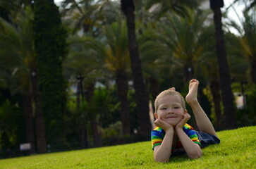 A little blond boy lies sitting on the bright green grass. The child in a multi-colored t-shirt with rainbow. Concept: Summer, family vacation, amusement park, entertainment, vacation. Space for text