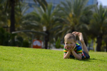 A little blond boy lies sitting on the bright green grass. The child in a multi-colored t-shirt with rainbow. Concept: Summer, family vacation, amusement park, entertainment, vacation. Space for text