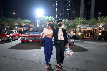 Lesbian couple with facial mask standing at a night street fair with vintage cars
