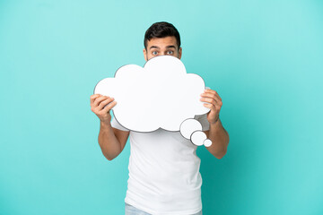 Young handsome caucasian man isolated on blue background holding a thinking speech bubble and hiding behind it